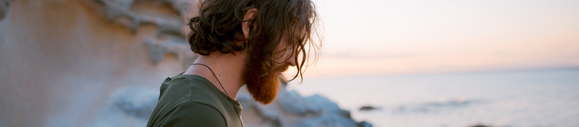 A bearded guy stood by the ocean looking down A bearded guy stood by the ocean looking down