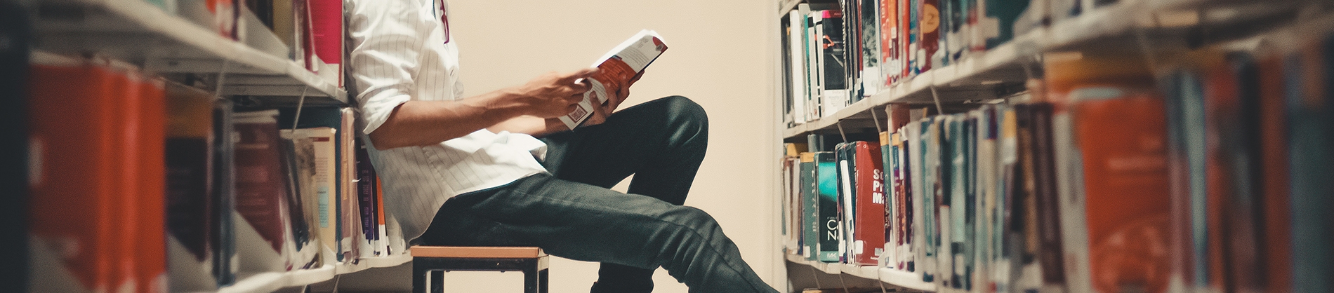 A man sat on a stool leaning on a book case reading A man sat on a stool leaning on a book case reading