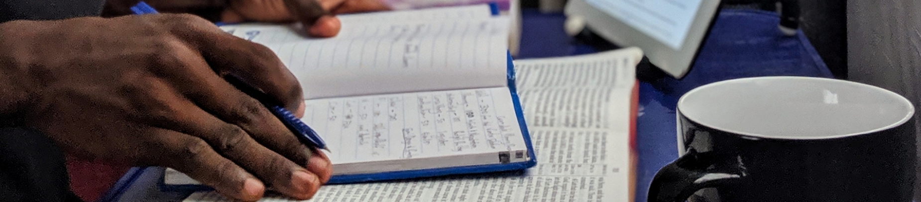 A man writing notes with his bible open and a mug beside him A man writing notes with his bible open and a mug beside him